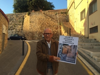 El veí de la comunitat, Josep Mir, mostra el cartell per demanar la instal·lació d’unes escales mecàniques i un ascensor per accedir a la plaça situada davant de la Diputació, ja que les escales actuals -al fons de la imatge- estan deteriorades